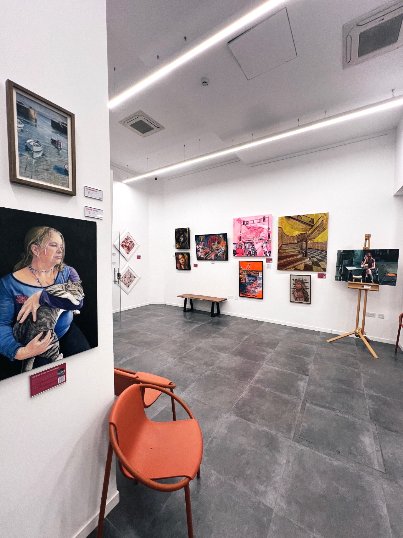 Bright exhibition room with paintings, an easel and orange chairs in the corner