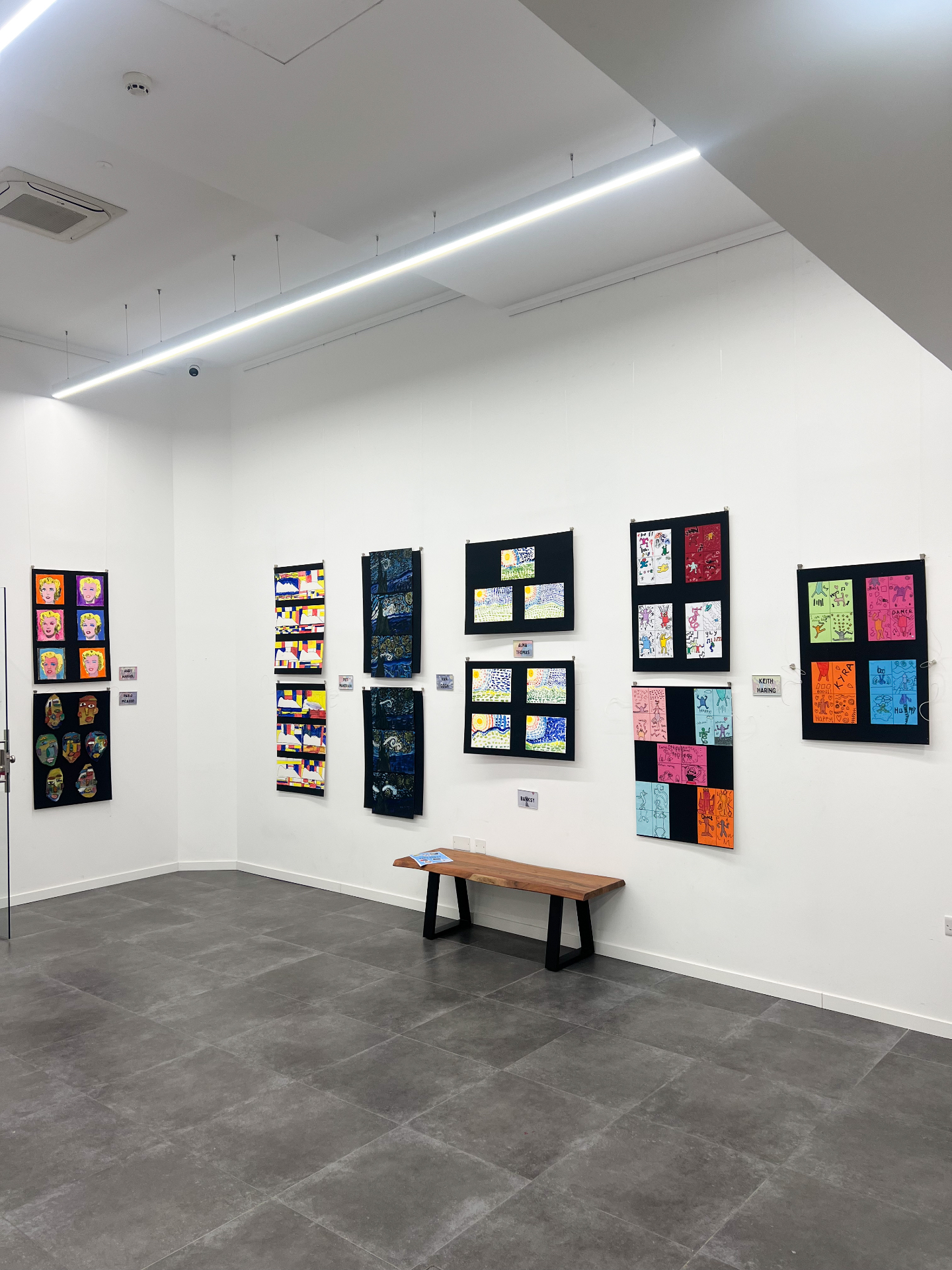 Gallery wall hung with colourful children's artwork in black frames above a wooden bench