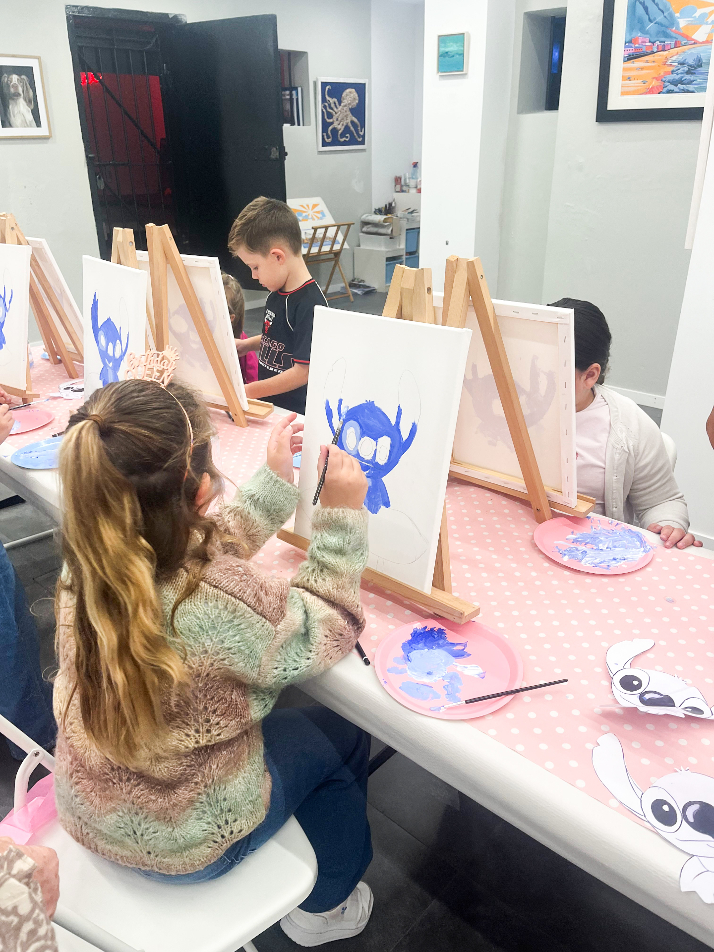 Children painting Stitch canvases at easels on a pink polka-dot party table