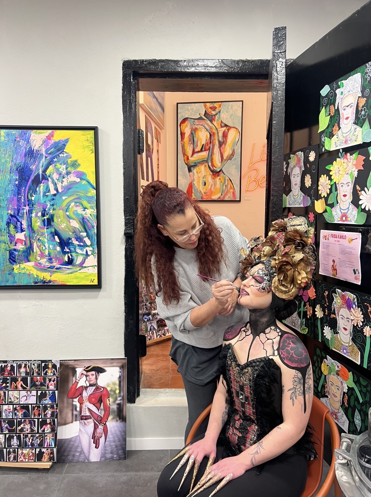 Artist applying body art to a model surrounded by paintings at the Women in Art Exhibition